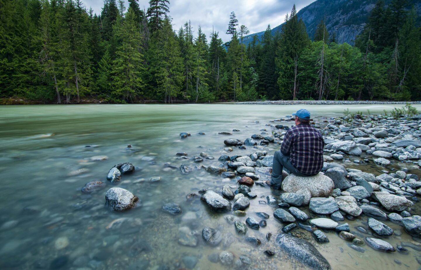Man sitting quietly on river rocks beside a forest stream, surrounded by nature—representing solitude, mindfulness, and the restorative power of slowing down during the recovery journey.