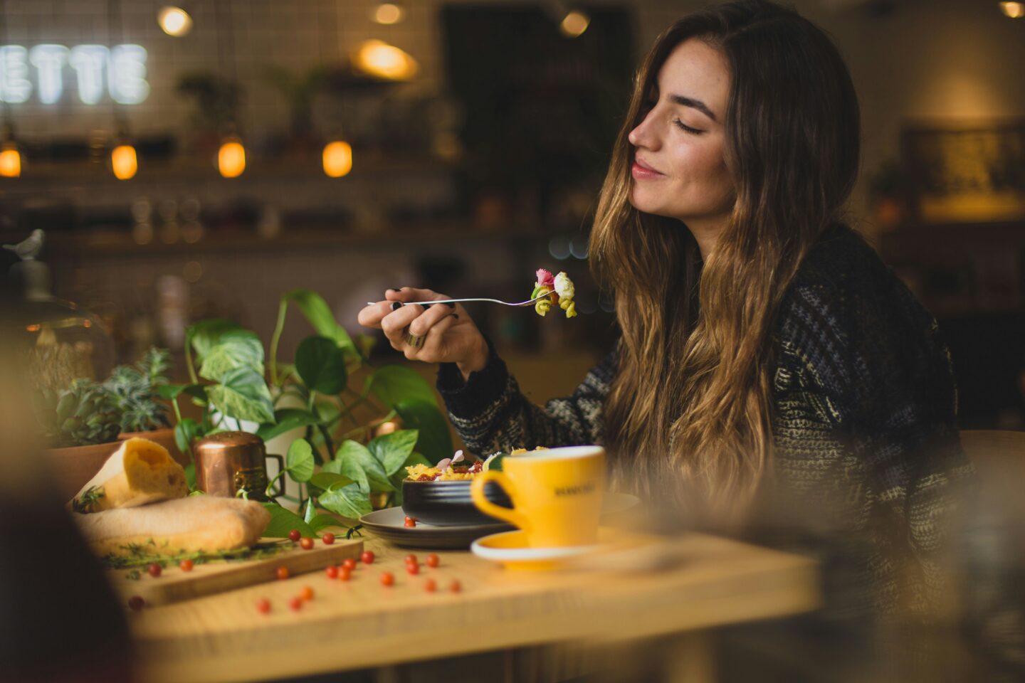 A woman smiling softly with eyes closed, savoring a bite of food at a cozy café table, surrounded by plants and vibrant dishes—capturing the essence of mindful eating and joyful nourishment promoted through DBT.