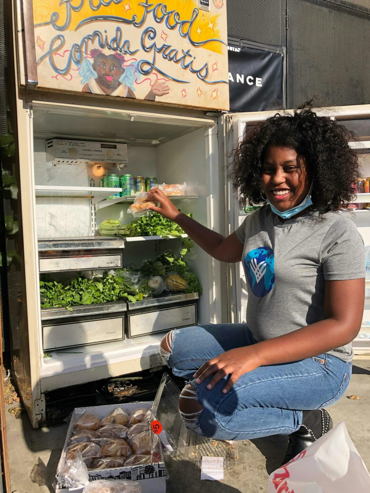Young Black woman smiling as she stocks a community fridge with fresh produce—highlighting food access, community care, and support for those facing economic barriers to recovery.