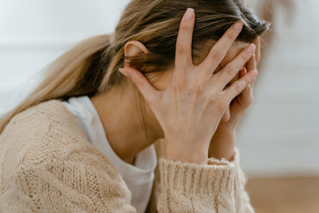 A woman sits with her head in her hands, overwhelmed or distressed. This image represents the emotional weight of body image struggles and the pressure to always feel positive about appearance.