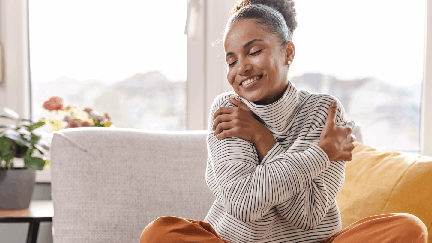 Woman sits criss-cross on a couch, smiling and hugging herself. Eating disorder recovery is possible. Take the first step by reaching out to Kindful Body in California today.