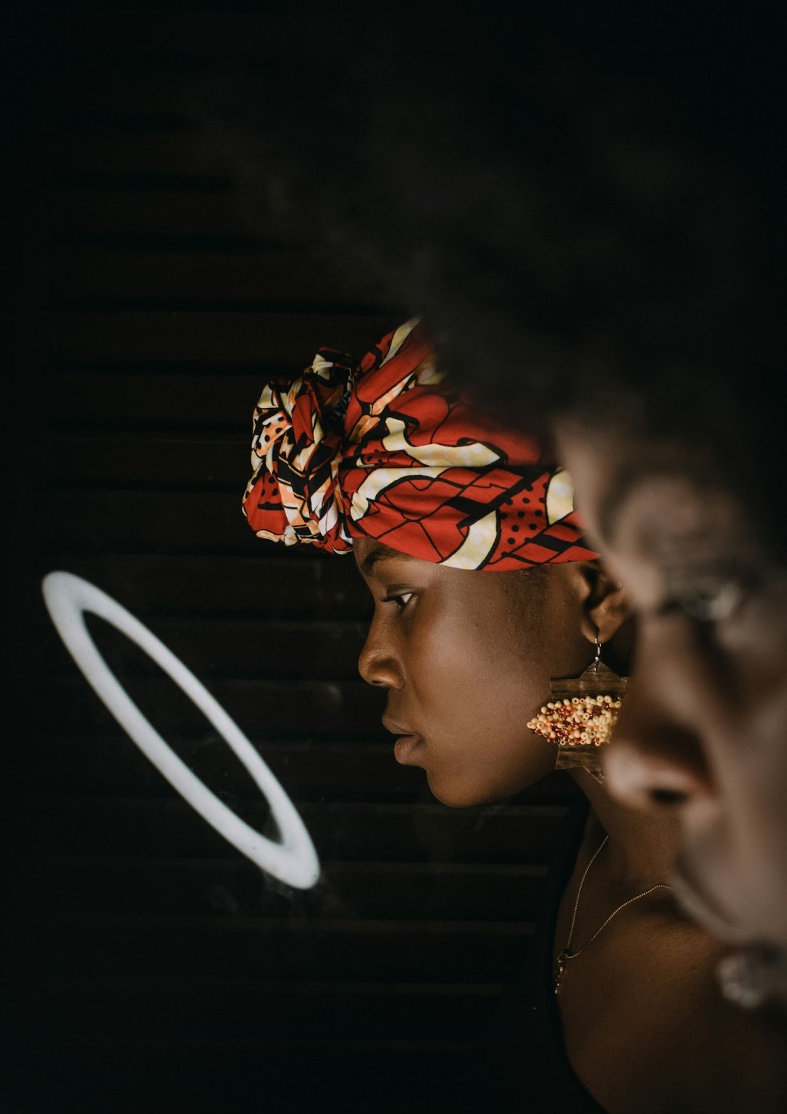 A woman wearing a vibrant red patterned headwrap looks into a circular mirror with a contemplative expression. The dark background and soft light emphasize themes of identity, beauty, and self-reflection within cultural and personal healing.