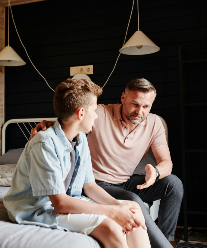 A father sits beside his son on a bed, speaking gently and offering support. The moment captures an open, caring conversation—symbolizing how parents can help children build healthy body image and self-esteem through empathy and listening.