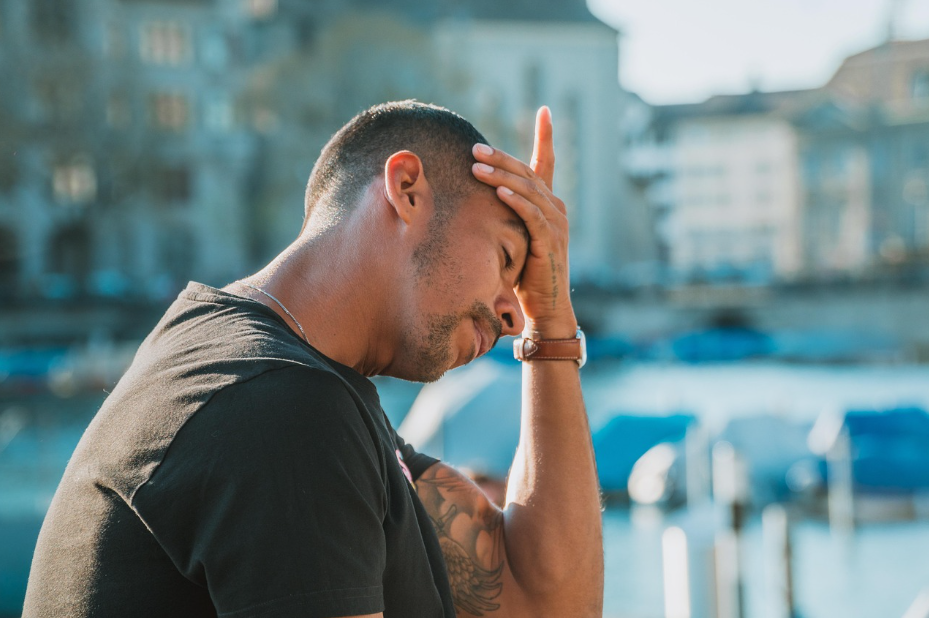 Person standing outside with eyes closed and hand on their forehead, showing stress or fatigue, with a bright city waterfront in the background.