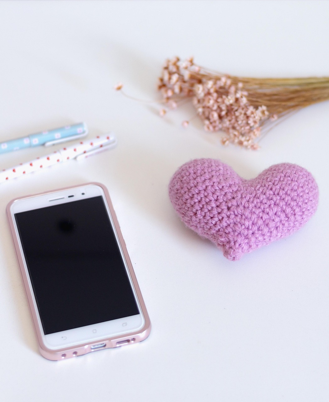 Smartphone on a white desk next to a handmade crocheted heart and dried flowers, symbolizing a gentler, more supportive online space.