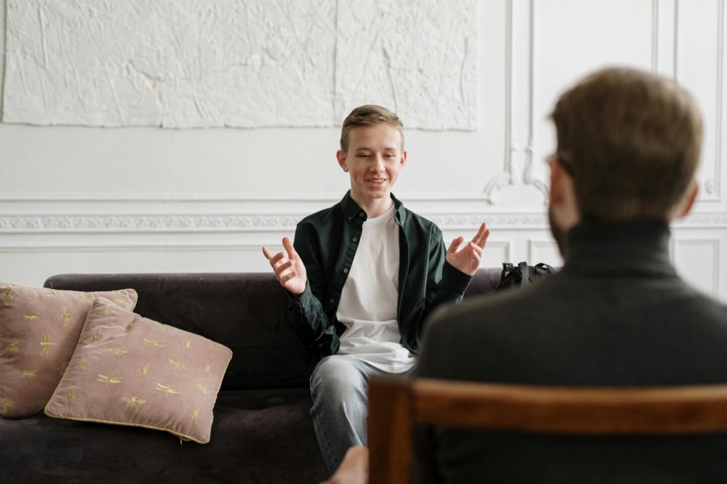 Therapist listening as a teen client talks and gestures while seated on a couch during a counseling session.