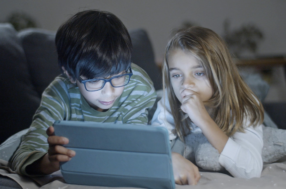Two young children lie on a couch watching a tablet together, focused and curious. The image reflects how kids absorb messages from media about appearance and identity, highlighting the importance of guiding early conversations about body image.